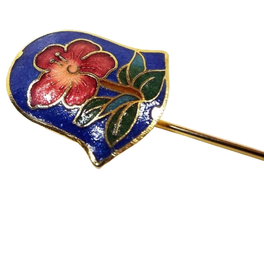 Magnified view of the top of the retro vintage cloisonne stick pin. The metal is gold tone in color and has a bell shape at the top. There is a pinkish red flower with green leaves on a dark blue enameled background.