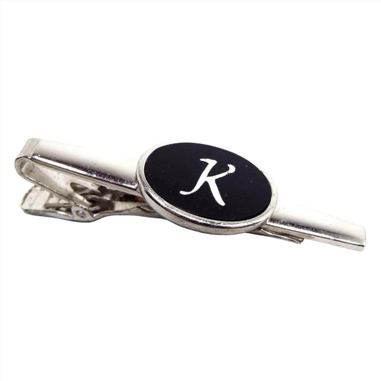 Angled top and side view of the Mid Century vintage Anson initial tie clip. It is silver tone in color. There is an oval on the front in the middle with a black plastic cab and the letter K etched in it. 