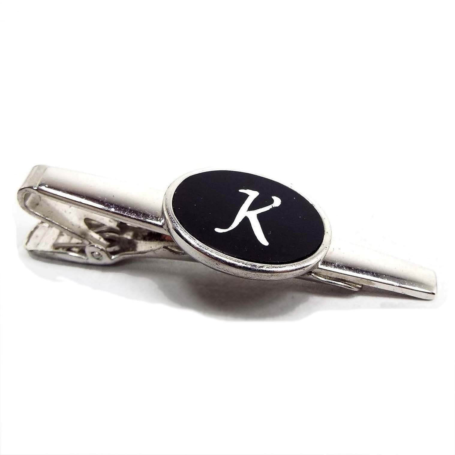 Angled top and side view of the Mid Century vintage Anson initial tie clip. It is silver tone in color. There is an oval on the front in the middle with a black plastic cab and the letter K etched in it. 