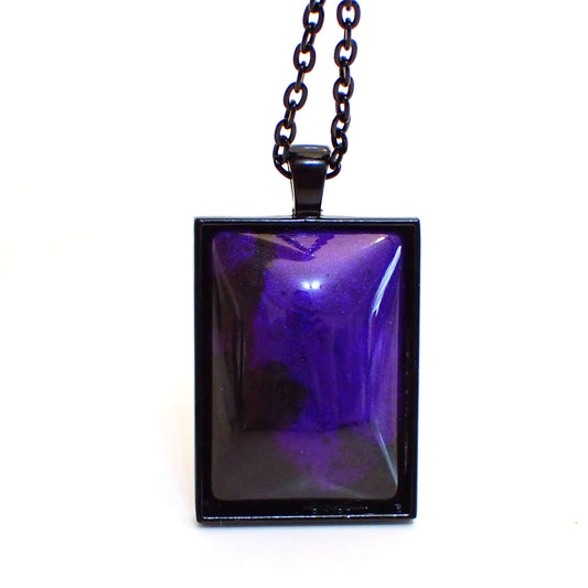 Enlarged front view of the large Goth rectangle pendant necklace. The chain and setting are black in color. The handmade resin cab is domed and has pearly black and purple resin. The purple resin has flashes of blue here and there depending on how the light hits it.