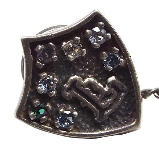 Front view of the Mid Century vintage initial tie tack. It is a very dark gray in color and is shaped like a shield. There is a fancy block letter L at the bottom middle and the top h as various tiny gemstone chips in different colors.