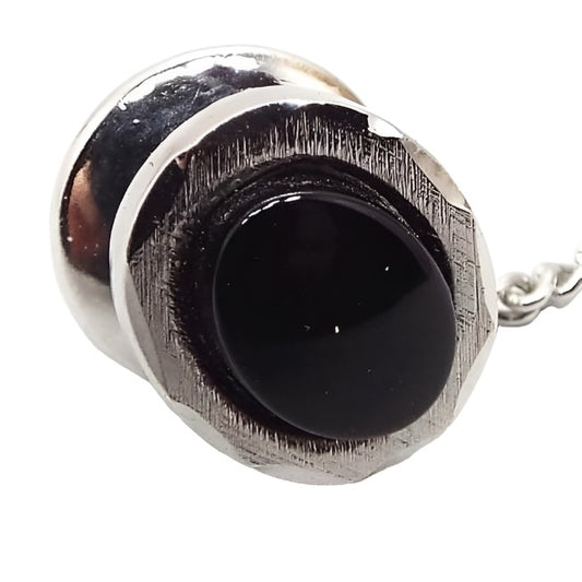 Front view of the retro vintage JL USA tie tack. It is faceted oval in shape with textured metal on the front. The sterling silver is slightly darkened from age. There is an oval black onyx gemstone cab in the middle.