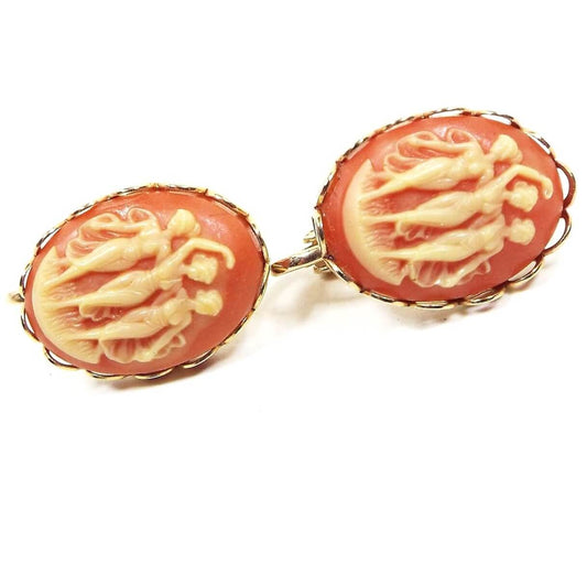 Front view of the retro vintage cameo clip on earrings. They have oval molded plastic cameos that are dark salmon pink in color with off white yellow depiction of "Three Graces" three women standing together. The metal is gold tone in color.