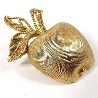 Front view of the retro vintage Avon apple brooch pin. It is gold tone in color and has a textured surface. It's shaped like an apple with stem and two leaves.