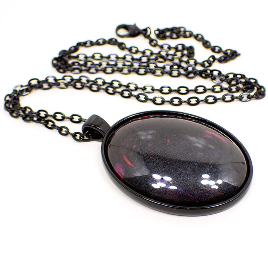 Angled view of the Goth large oval pendant necklace with handmade resin cab. The chain and setting are black in color. The chain has a lobster claw clasp. There is a large oval pendant at the bottom with a resin cab that is mostly pearly black with just a few small hints of red, blue, purple, and pink.