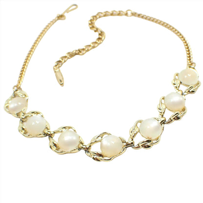 Angled front view of the Mid Century vintage link necklace. The metal is gold tone in color. There is a chain on either side with a hook clasp at one end. The bottom part of the necklace has open links with leaf like areas on one side of each link. There are domed round off white moonglow lucite cabs on each link. The cabs have an inner glowy effect as you move around in the light.