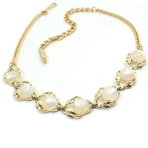 Angled front view of the Mid Century vintage link necklace. The metal is gold tone in color. There is a chain on either side with a hook clasp at one end. The bottom part of the necklace has open links with leaf like areas on one side of each link. There are domed round off white moonglow lucite cabs on each link. The cabs have an inner glowy effect as you move around in the light.