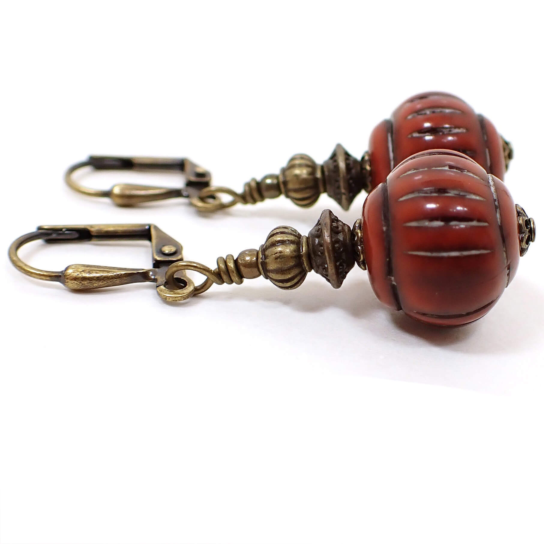 Side view of the handmade drop earrings. The metal is antiqued brass in color. There are lantern shaped lucite beads on the bottom that have a carved like line design. The beads are a dark cinnamon red in color.