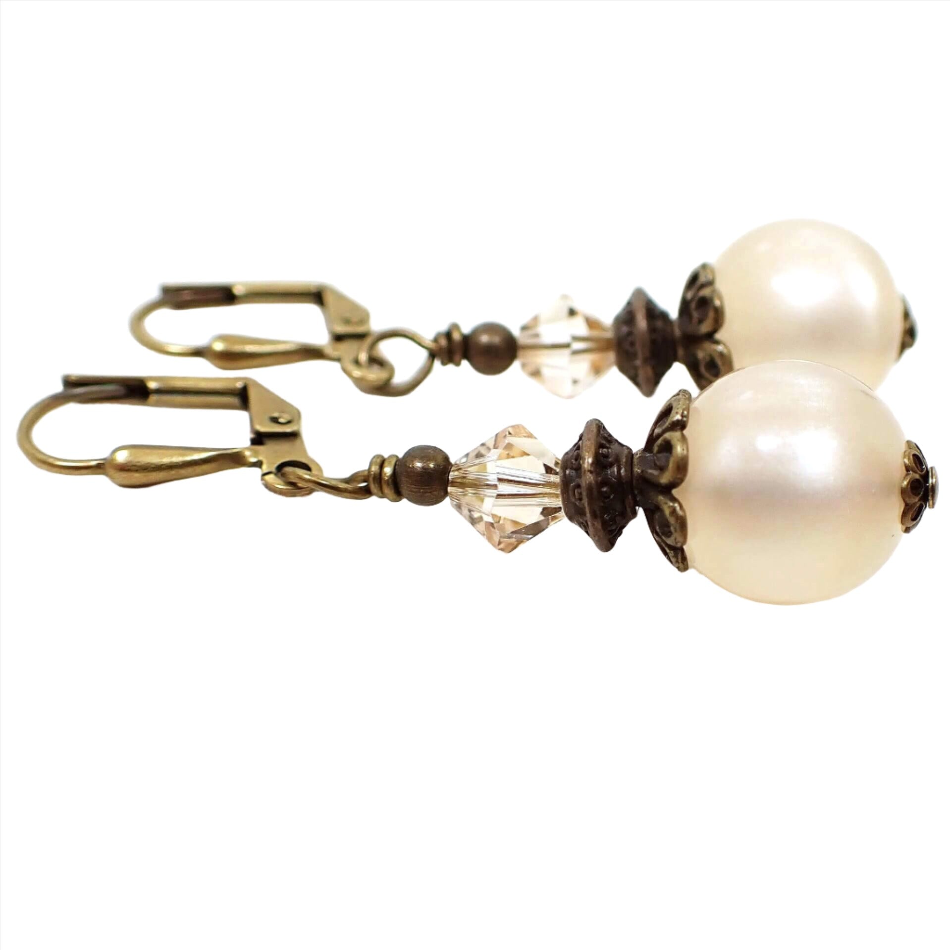 Side view of the handmade drop earrings with vintage lucite beads. The metal is antiqued brass in color. There are faceted glass bicone beads at the top in a very light champagne color. The bottom beads are round moonglow lucite in an off white color. They have a glowy effect as you move around in the light.