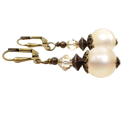 Side view of the handmade drop earrings with vintage lucite beads. The metal is antiqued brass in color. There are faceted glass bicone beads at the top in a very light champagne color. The bottom beads are round moonglow lucite in an off white color. They have a glowy effect as you move around in the light.