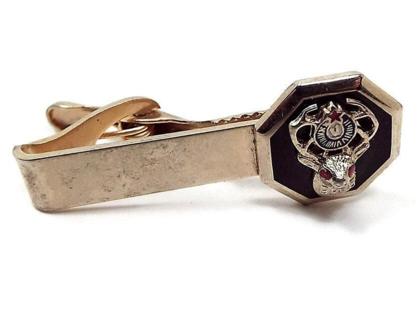 Front view of the Mid Century vintage Elks Tie Clip. The metal is gold tone in color. There is an octagon shape at the end of the tie clip with the fraternal Elks symbol of an elk with star and BPOE on it. The star and elk's eyes are enameled red and the background is enameled black.