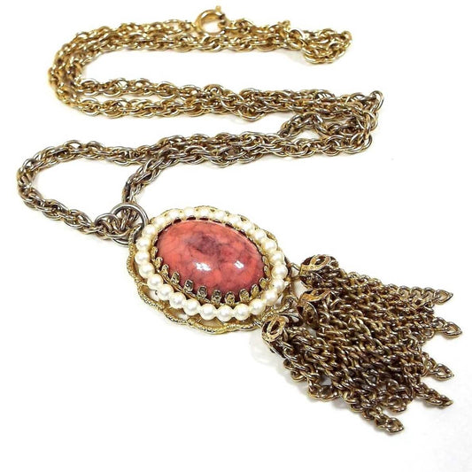 Front view of the retro vintage tassel necklace. The metal is a darkened gold tone in color. The pendant at the bottom is oval with a dark pink and gray marbled faux stone cab and faux pearls around the edge. There are three bunches of chain tassels at the bottom.
