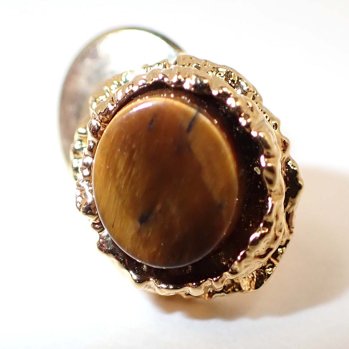 Front view of the Mid Century vintage Brutalist style tie tack. The metal is gold tone in color. There is a flat round tiger's eye gemstone cab in the middle. The surrounding setting has a free form bumpy textured style look. 