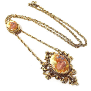 Front view of the Mid Century vintage cameo pendant necklace. The metal is antiqued gold tone in color. There is a long rope chain down to an oval cameo pendant of two people sitting together. The chain extends down past that to another filigree shield shaped pendant with another oval cameo cab and small off white faux pearls around the top area. The cameos are smooth plastic with decals over them.