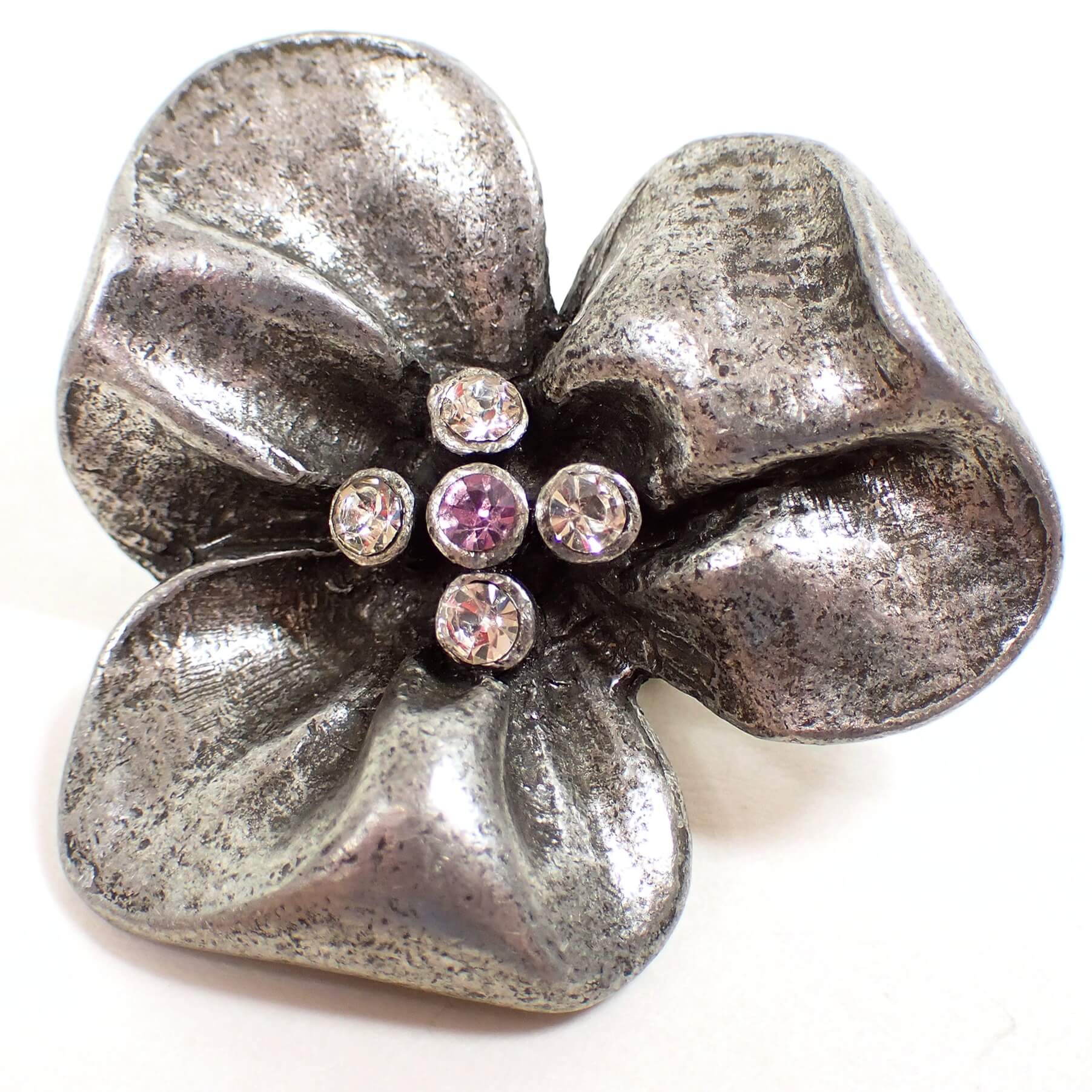 Front view of the retro vintage flower broch pin. The metal is pewter and has a gray and dark gray appearance. There are three rounded wide petals with rhinestones in the middle of the flower. There are four small round clear rhinestones surrounding a middle rhinestone that is pink or purple depending on the lighting.