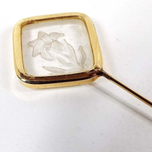 Enlarged top view of the retro vintage intaglio glass floral stick pin. The metal is gold tone in color. There is a diamond shape at the top with a glass insert. The inserts has a flower etched into the back of it to give a raised appearance on the front.