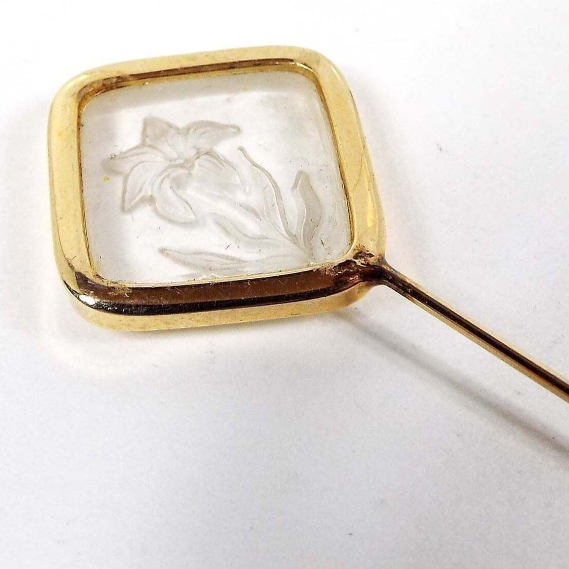 Enlarged top view of the retro vintage intaglio glass floral stick pin. The metal is gold tone in color. There is a diamond shape at the top with a glass insert. The inserts has a flower etched into the back of it to give a raised appearance on the front.