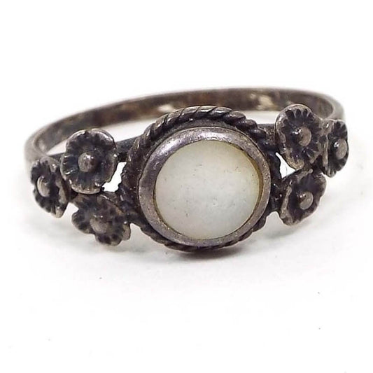 Front view of the retro vintage sterling silver and mother of pearl flower ring. The sterling is very darkened to a gray color. In the middle is a bezel set pearly white mother of pearl shell flat cab with a braided rope style edge around it. On each side is a set of three small sterling flowers. 