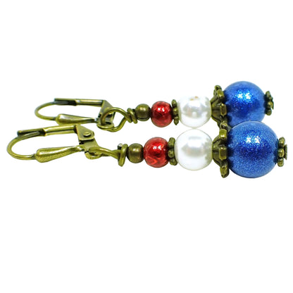 Enlarged side view of the red white and blue handmade earrings. The metal is antiqued brass in color. There is a small metallic red glass bead at the top, a white faux pearl bead in the middle, and a sparkly blue bead at the bottom.