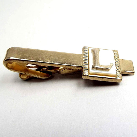 Angled top view of the Mid Century vintage initial tie clip. It is gold tone in color. There is a pearly white mother of pearl cab at the end with the letter L on it. 