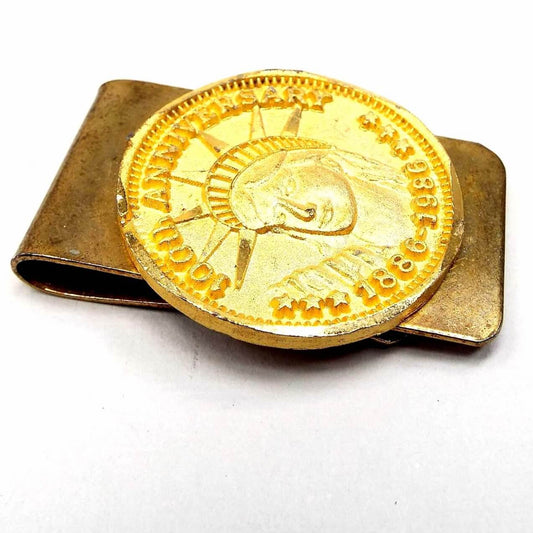 Angled top view of the retro vintage faux coin money clip. The clip base is a darkened gold tone in color from age. On the front is a bright gold tone color imitation coin that has the Statue of Liberty head and the words 100th Anniversary 1886-1986. The edges of the faux coin are faceted.