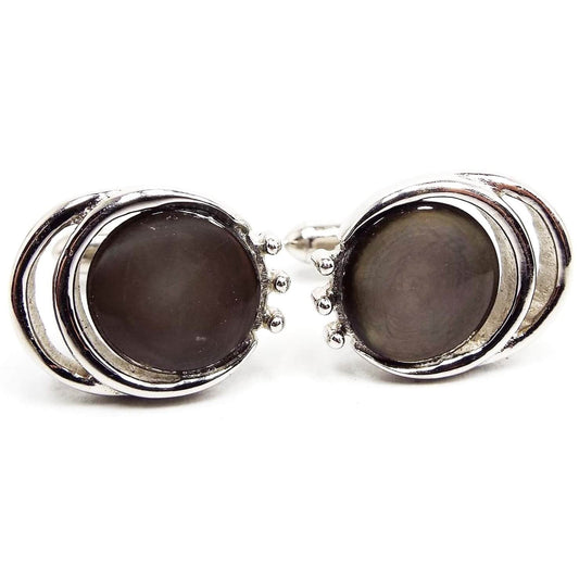 Front view of the Mid Century vintage Swank cufflinks. They are round with two cut out rounded edges on the side. There is a round mother of pearl cab that is dyed a brownish gray color. The metal is silver tone in color.