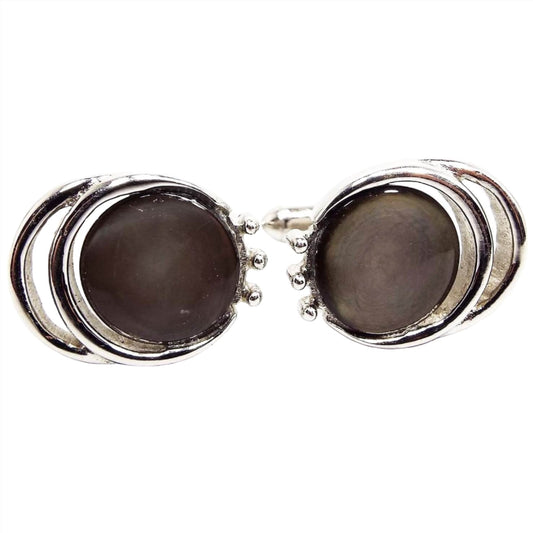Front view of the Mid Century vintage Swank cufflinks. They are round with two cut out rounded edges on the side. There is a round mother of pearl cab that is dyed a brownish gray color. The metal is silver tone in color.