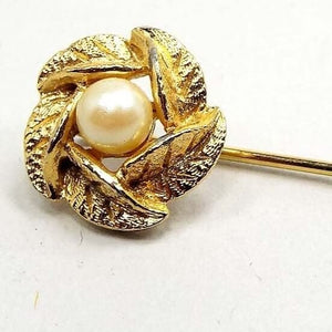 Enlarged top view of the Mid Century vintage faux pearl stick pin. The metal is gold tone in color. The top has a wreath style design made of textured metal leaves. There is an off white imitation pearl in the middle.
