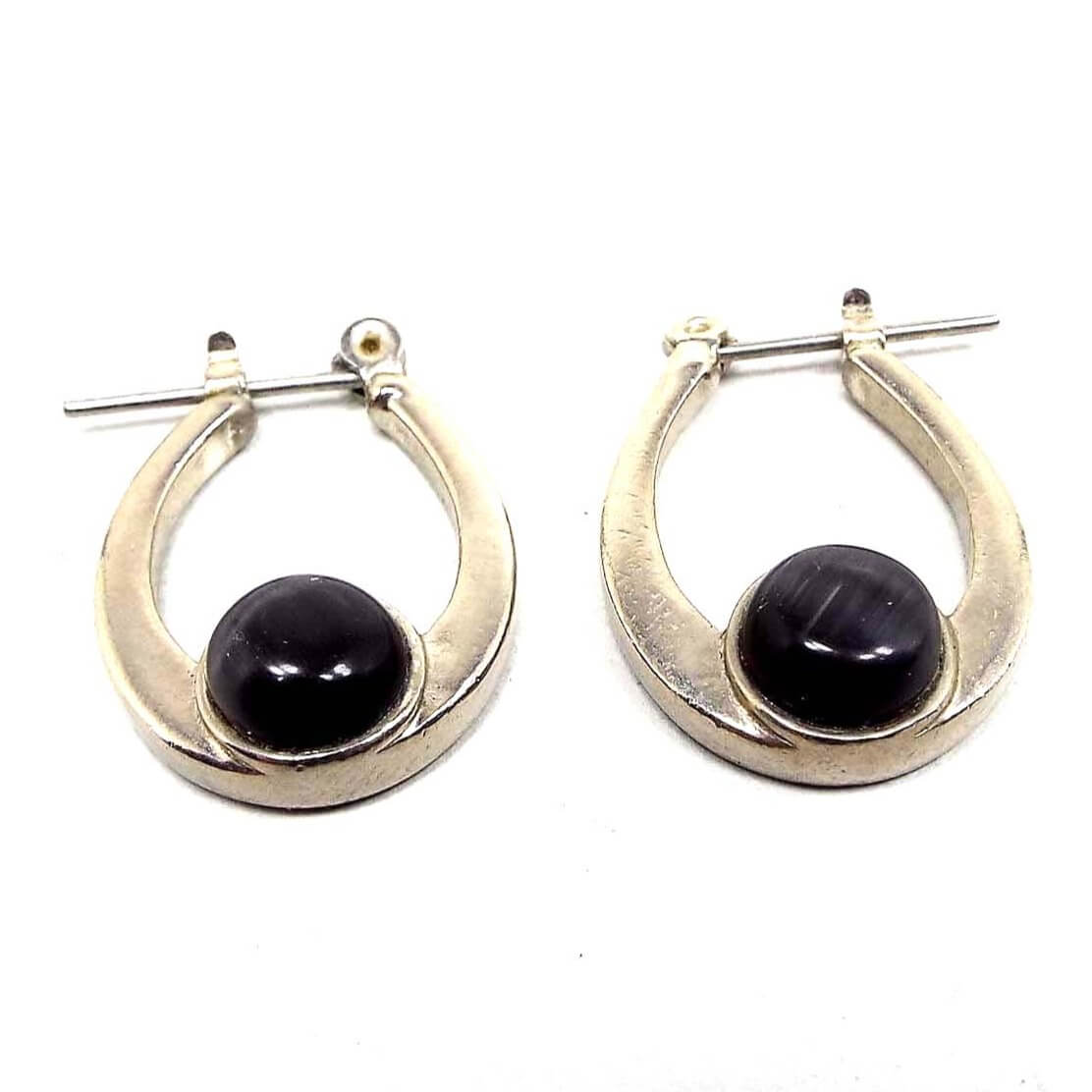 Angled side view of the retro vintage glass faux cat's eye hoop earrings. The metal is silver tone in color. The hoops are oval teardrop shaped. At the bottom is a glass cab in dark gray that has flashes of lighter gray as you move around in the light. Earrings are one sided.
