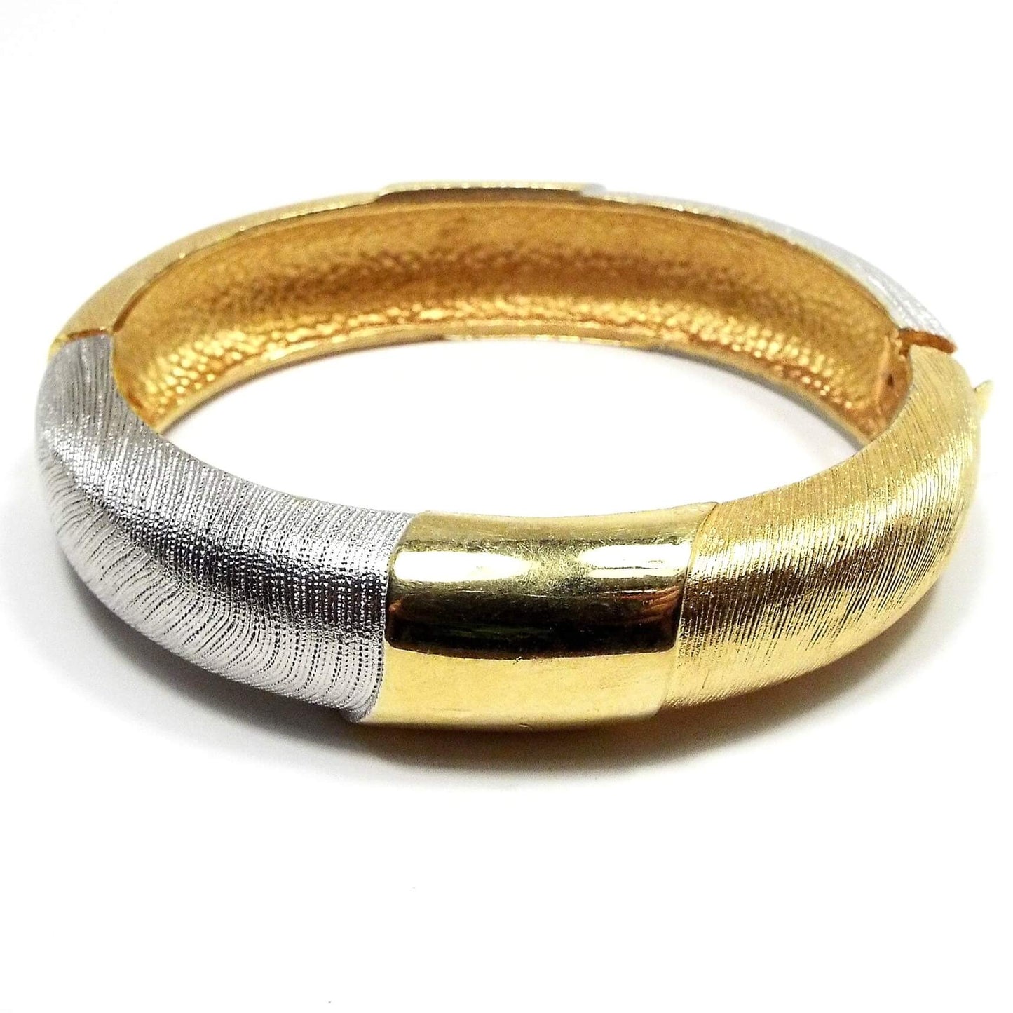 Angled side view of the retro vintage hinged bangle bracelet. It is two tone in color with gold tone and silver tone color metal. Most of the bangle has a textured line design on it. There is a locking hinge clasp at the side.