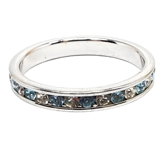 Angled view of the retro vintage rhinestone eternity band ring. It is a thinner style band with silver tone color metal. There is a channel that goes all the way around the band and has alternating light blue and clear rhinestones in it.