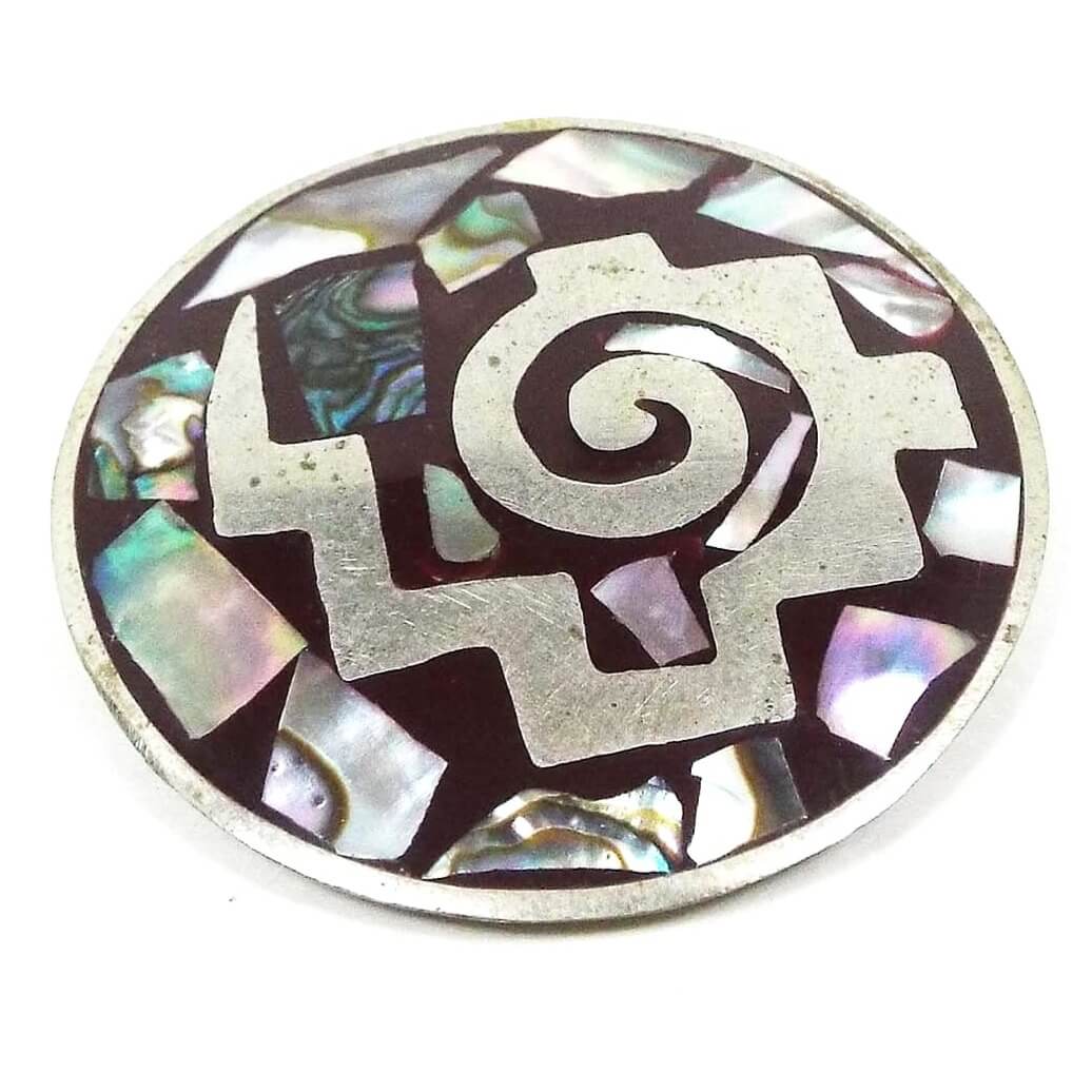 Front view of the retro vintage Mexican enameled brooch pendant. It is round in shape with silver tone color metal. It is dark brown enameled with a silver tone shape that looks like a swirl with steps on the outside and a lightning bolt shaped tail. There are irregularly shaped pieces of pearly multi color abalone shell around the rest of the brooch with dark brown enamel surrounding them.