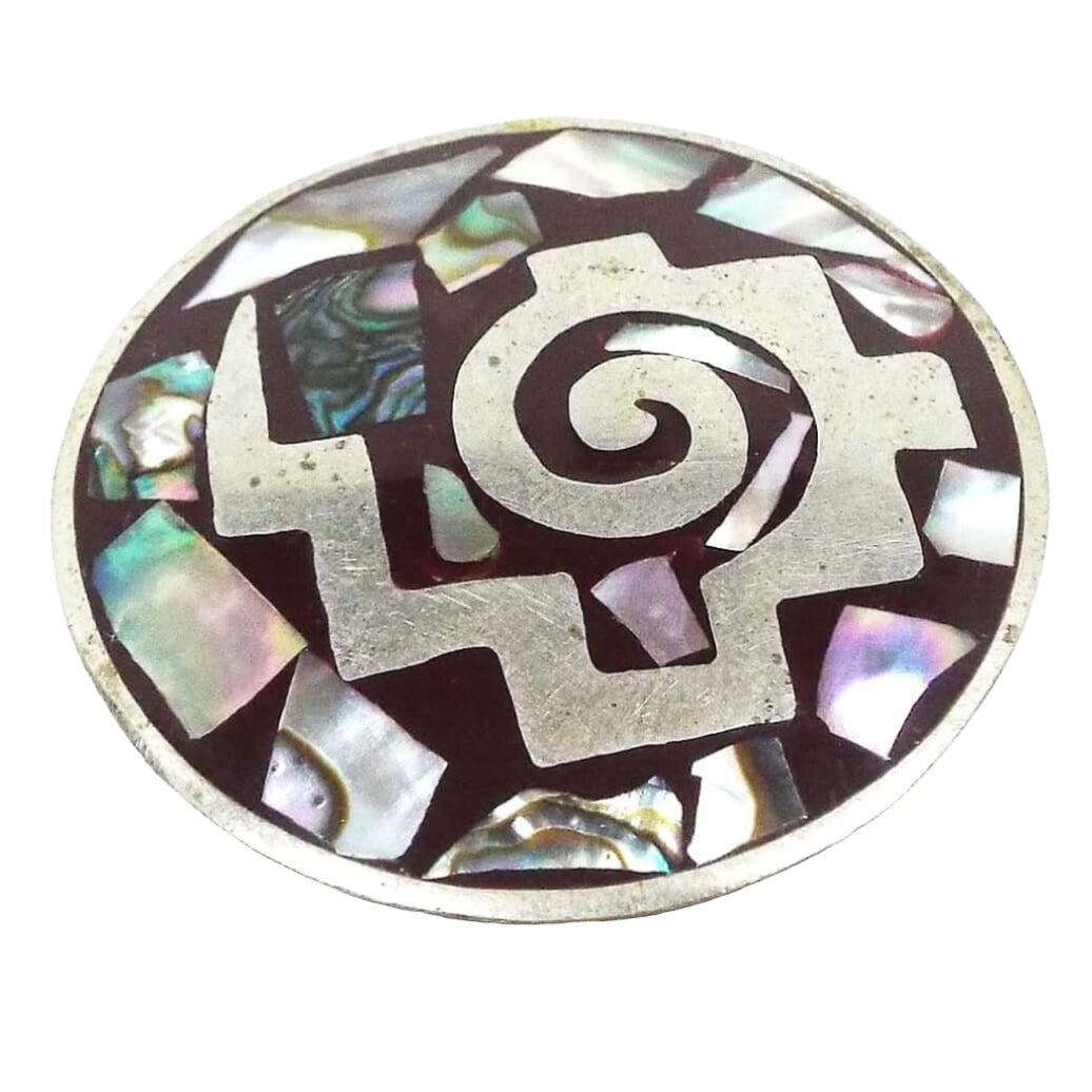 Front view of the retro vintage Mexican enameled brooch pendant. It is round in shape with silver tone color metal. It is dark brown enameled with a silver tone shape that looks like a swirl with steps on the outside and a lightning bolt shaped tail. There are irregularly shaped pieces of pearly multi color abalone shell around the rest of the brooch with dark brown enamel surrounding them.