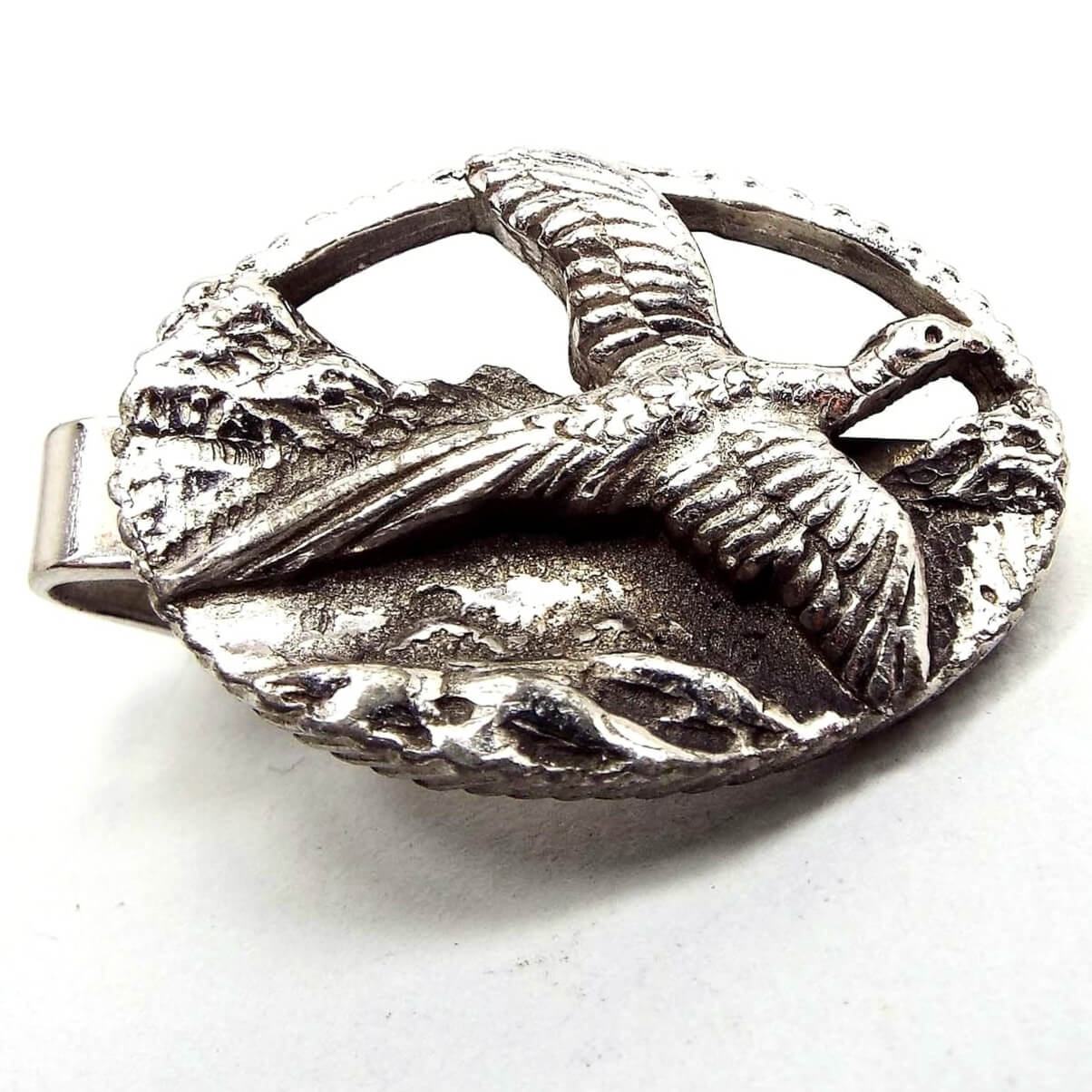 Front view of the retro vintage pheasant tie clip. It is silver tone and oval in shape. There is a raised and detailed design of a pheasant flying with what looks to be mountains in the background.
