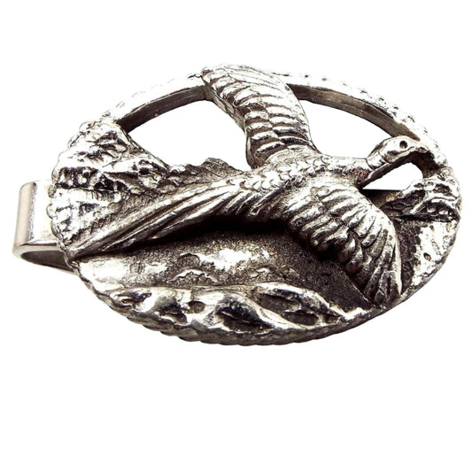 Front view of the retro vintage pheasant tie clip. It is silver tone and oval in shape. There is a raised and detailed design of a pheasant flying with what looks to be mountains in the background.