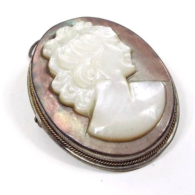Angled front view of the 1940's vintage cameo brooch pendant. It is oval in shape with mostly a silver tone color bezel setting that has a thin twisted style gold tone trim around the edge. The middle is a carved mother of pearl cab. The middle part is a pearly white bust of a woman and the background is a pearly brownish gray in color.
