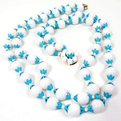 The Mid Century vintage Hong Kong multi strand beaded necklace. It has two strands of large round white plastic beads and blue enameled aluminum bead caps in a flower like design. There is a round beaded box clasp at the end.