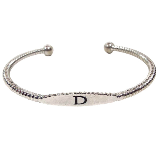 Angled front view of the retro vintage initial cuff bracelet. The metal is silver tone in color. There is a flat oval area on the front with the letter D on it. The rest of the bracelet has two thick rounded  wires that go back to ball ends. The wires have a twist like design. 