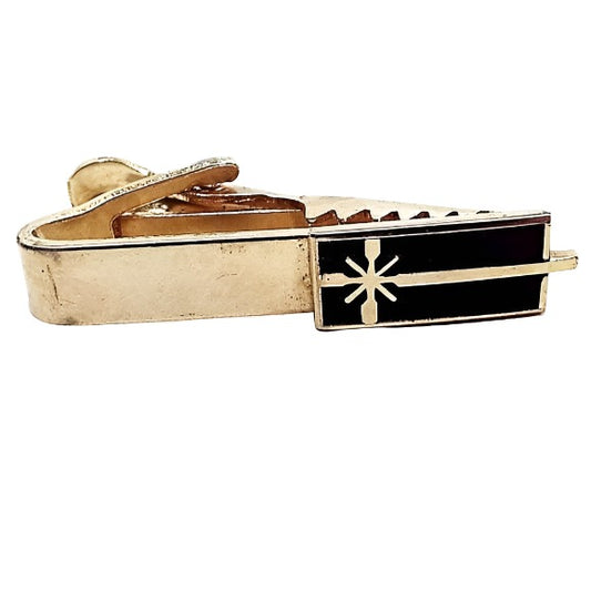 Front view of the Mid Century vintage enameled tie clip. It is gold tone in color and has a black enameled rectangle on the end with a gold tone starburst design One of the lines of the starburst extends to the end of the tie clip. 