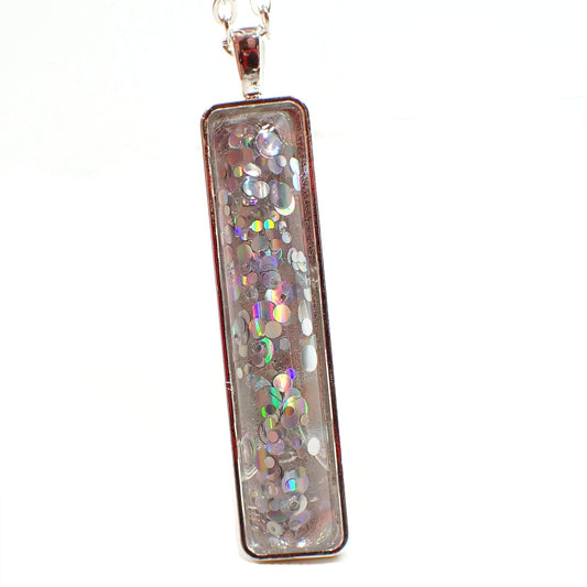 Front view of the handmade glitter pendant. The metal is silver tone in color. There is a long clear rectangle resin cab on the front of the pendant that has round bits of holographic glitter in it. The glitter has flashes of rainbow colors as you move around in the light.