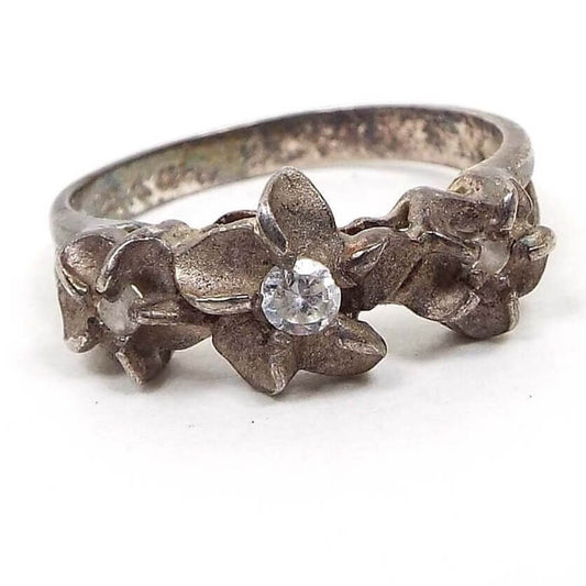 Front view of the Mid Century vintage sterling silver flower ring with rhinestones. The silver is very darkened to a gray color from age. There are three flowers at the top of the ring with matte sterling petals and small prong set round rhinestones in the middle. 