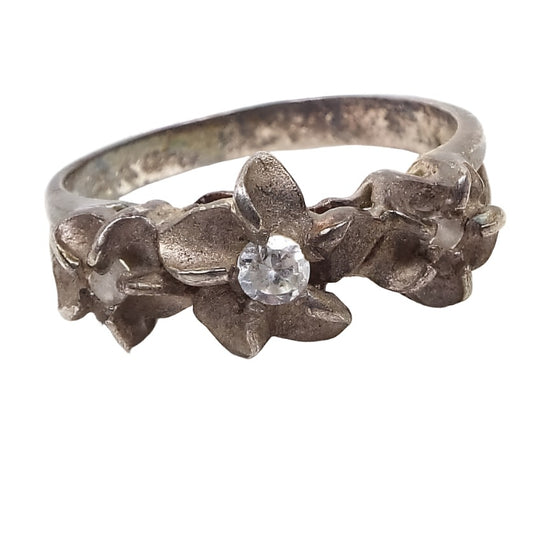 Front view of the Mid Century vintage sterling silver flower ring with rhinestones. The silver is very darkened to a gray color from age. There are three flowers at the top of the ring with matte sterling petals and small prong set round rhinestones in the middle. 