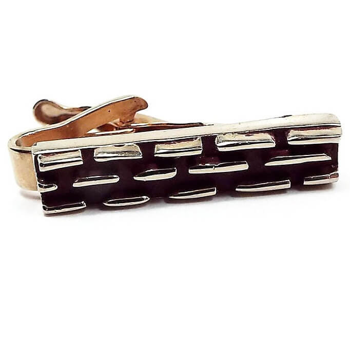 Front view of the smaller sized retro vintage tie clip. It is gold tone in color with a raised line dash style design. The background is painted a dark brown color.
