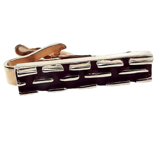 Front view of the smaller sized retro vintage tie clip. It is gold tone in color with a raised line dash style design. The background is painted a dark brown color.