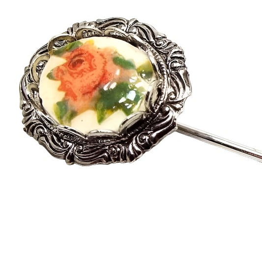 Enlarged view of the top of the Mid Century vintage floral stick pin. The metal is silver tone in color. There is a textured oval frame at the top with an oval plastic cab. The cab has a rose flower decal with shades of pink and green.