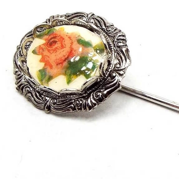 Enlarged view of the top of the Mid Century vintage floral stick pin. The metal is silver tone in color. There is a textured oval frame at the top with an oval plastic cab. The cab has a rose flower decal with shades of pink and green.