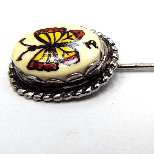 Enlarged view of the top of the retro vintage butterfly stick pin. The metal is darker silver tone in color. The top has an oval scalloped bezel with an off white glass cab. On the front of the cab is a yellow and reddish orange painted butterfly. The artists signature symbol is painted under the butterfly.