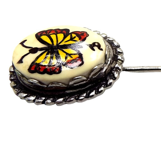 Enlarged view of the top of the retro vintage butterfly stick pin. The metal is darker silver tone in color. The top has an oval scalloped bezel with an off white glass cab. On the front of the cab is a yellow and reddish orange painted butterfly. The artists signature symbol is painted under the butterfly.