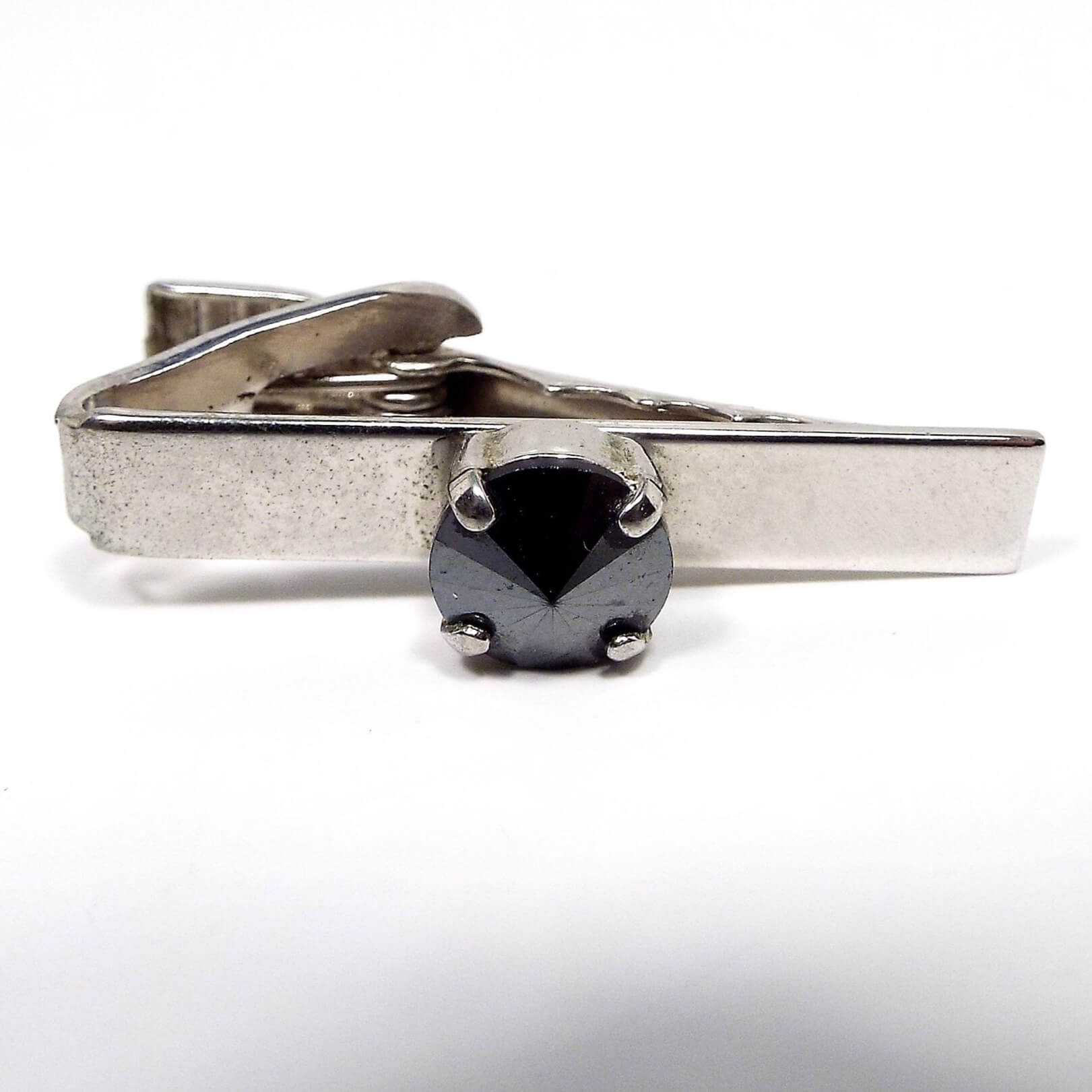 Front view of the Mid Century vintage faux hematite tie clip. The metal is silver tone in color. In the middle is a prong set rivoili rhinestone that is dark metallic gray in color.