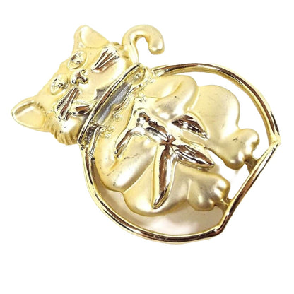 Front view of the AJC retro vintage cat in fish bowl brooch pin. The metal is matte gold tone in color except for the fish bowl which is shiny. The cat is sitting in the fish bowl with his head and tail out and he's holding the goldfish in the bowl.